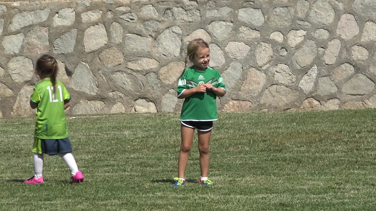A blond child in a green jersey stands near center on the grass in San Jose del Cabo, Mexico, as another child in a lime jersey passes in front of the stone wall.