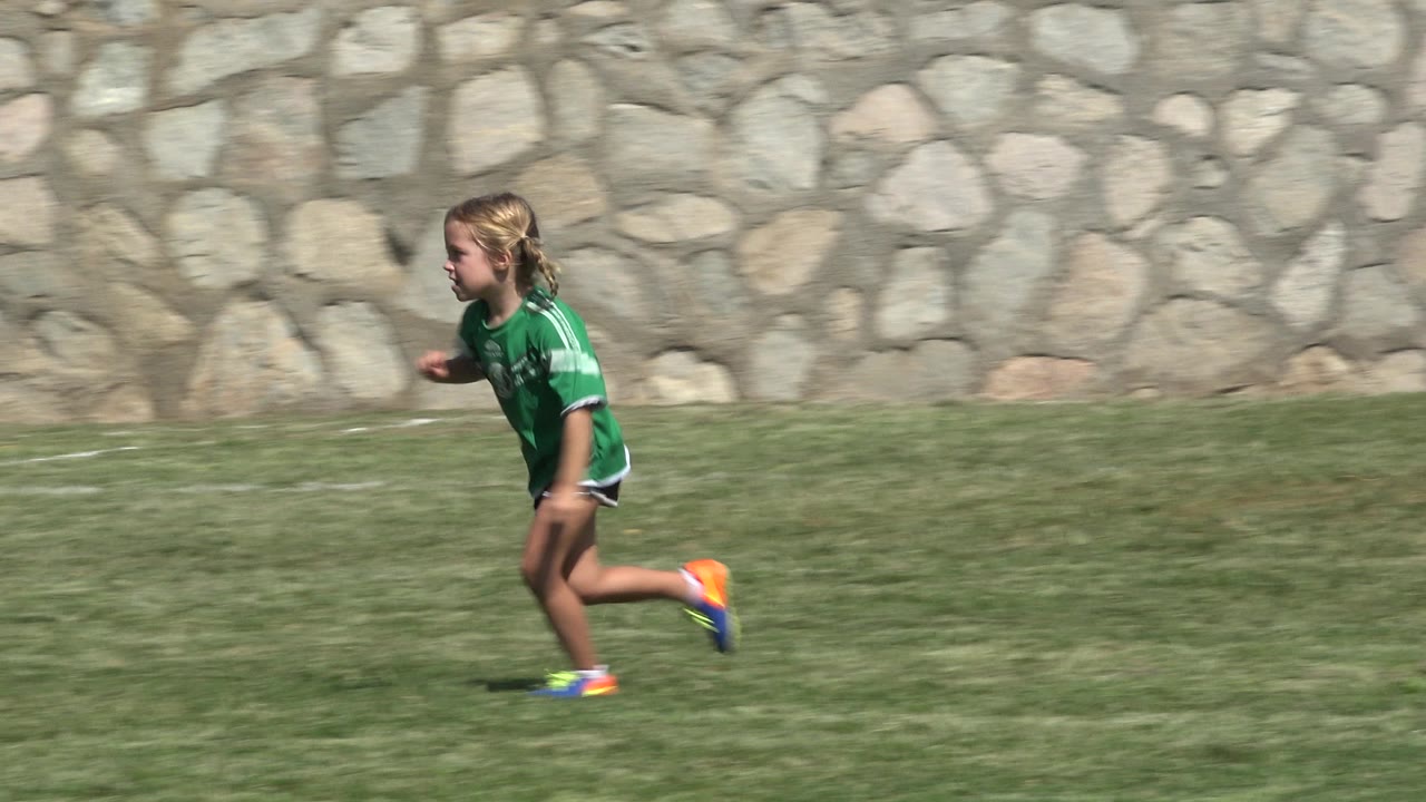 Blonde child in a green soccer jersey runs across a grass field.
