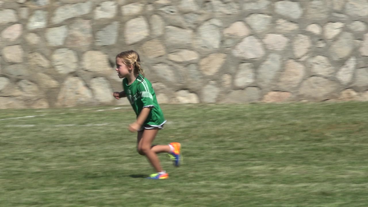 The same young player in a green jersey continues running in profile across the grass field, with the stone wall and bright midday light still visible.