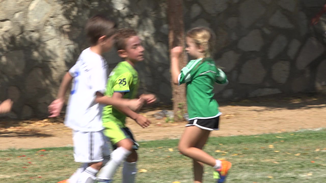 Children in white and green soccer uniforms run beside a stone wall.