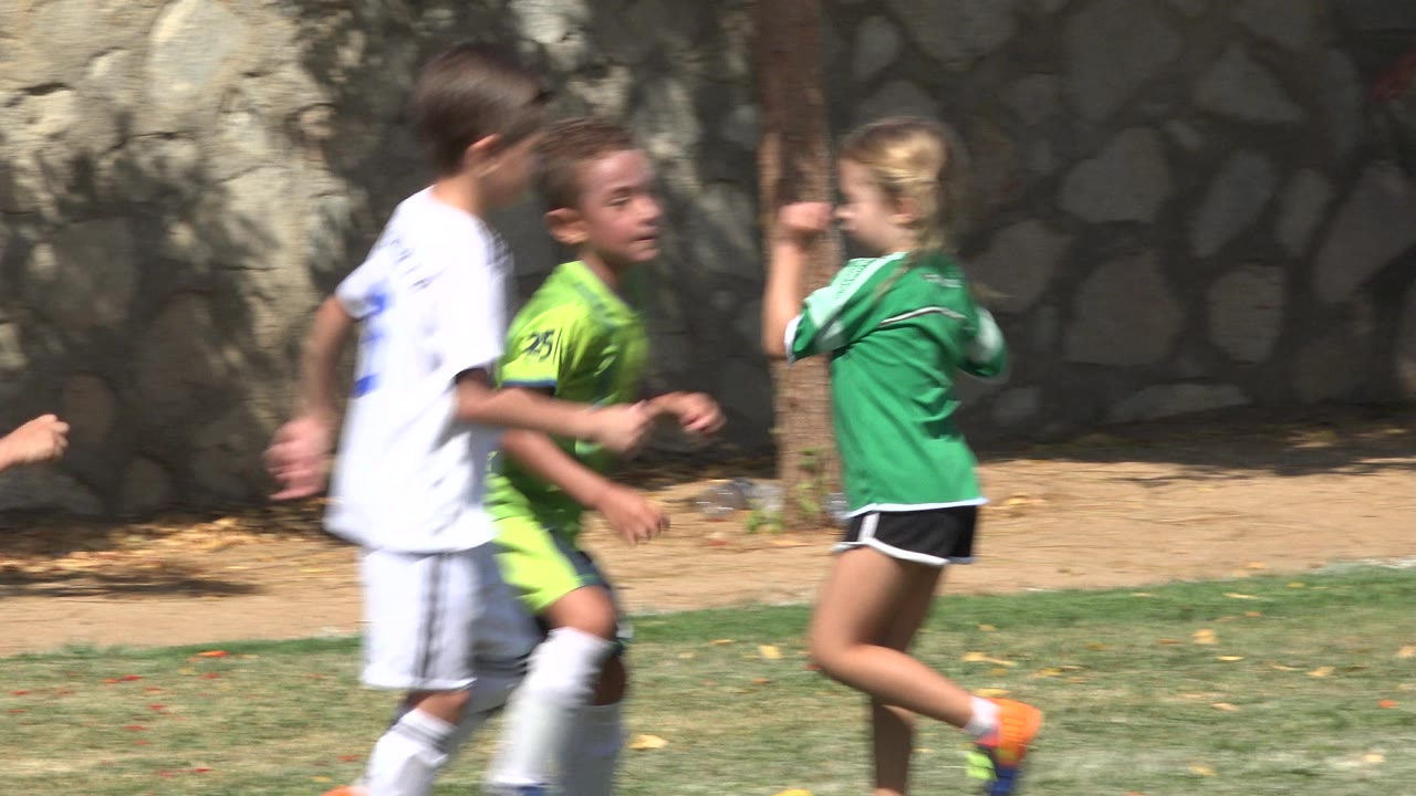 Three young players in white and green uniforms move through the frame in San Jose del Cabo, Mexico, while a stone wall and shaded dirt path line the background.