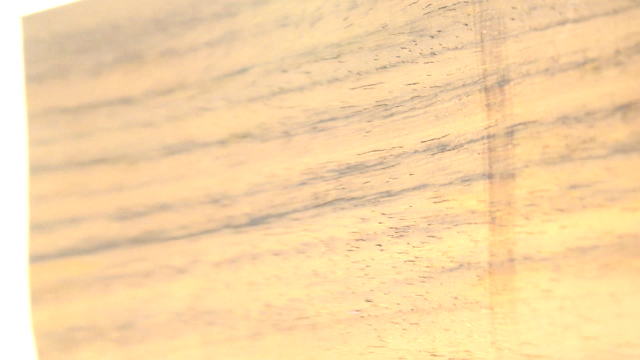 At 0:01, the wood slice remains close to camera with soft tan grain, a narrow glowing white strip on the left, and no visible people.