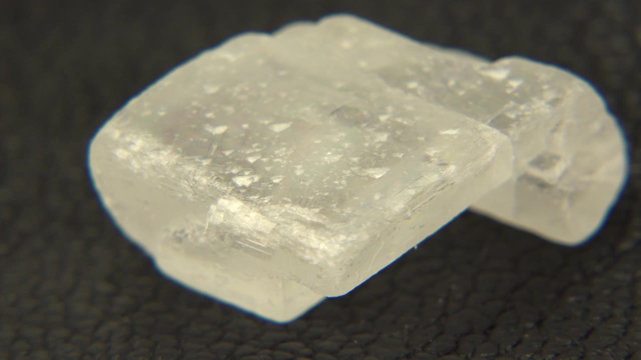 The white quartz crystal remains close to camera on the dark surface, showing pale flecks, translucent planes, and no visible people.