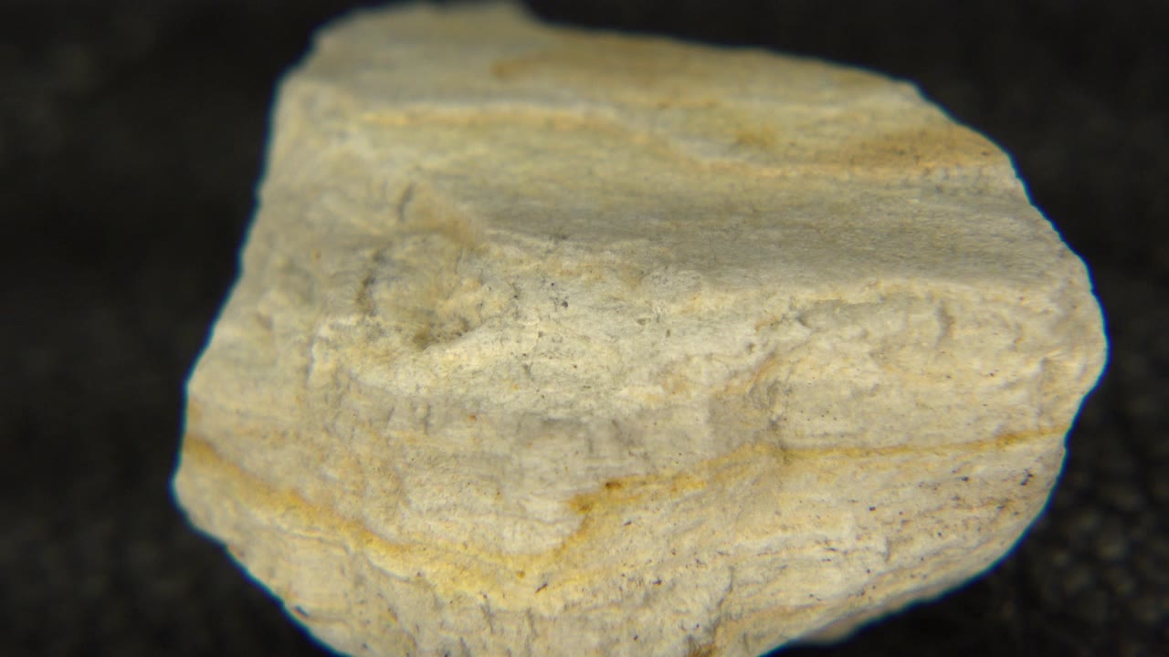 At 0:01, the white-gray rock has rotated slightly, showing a flatter upper face and a rough front edge on the dark table in Westminster, Colorado, with no visible people.