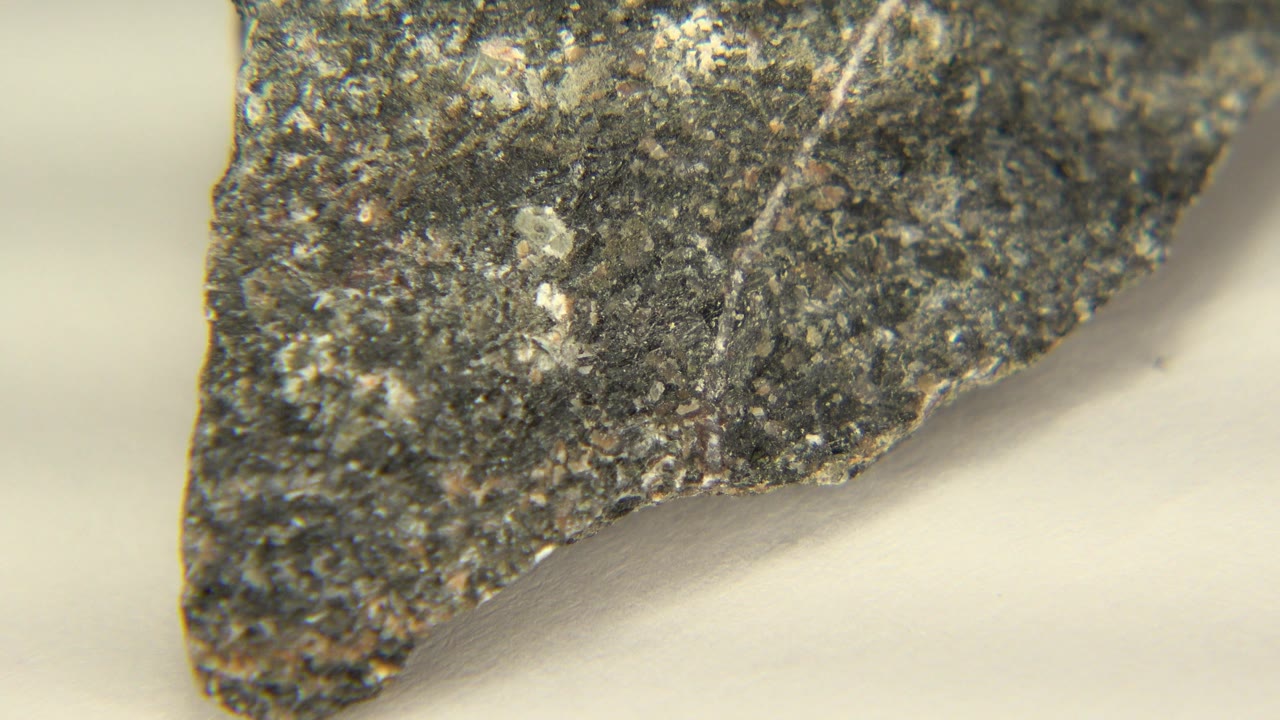 At 0:01, the rock remains close in frame with a pointed lower edge, rough gray texture, and scattered white mineral flecks, with no visible people.