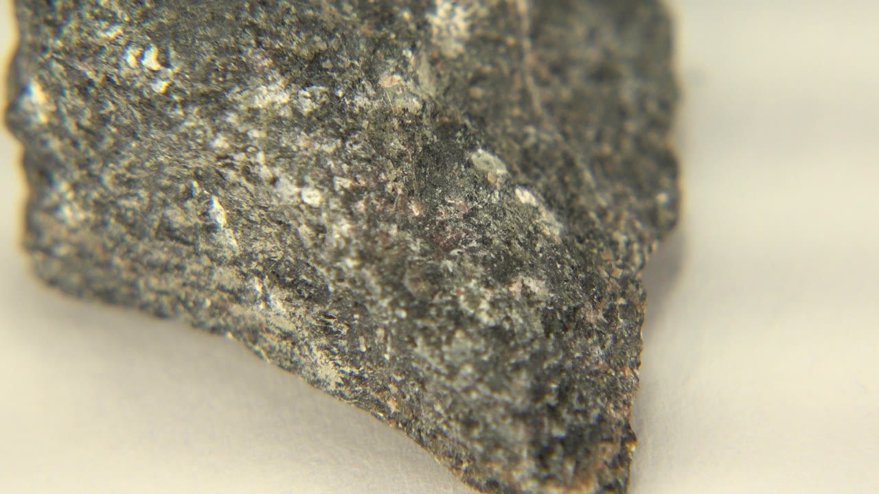 The dark rock remains close to camera on the neutral tabletop, showing a sharper pointed edge and flecked gray surface, with no visible people.