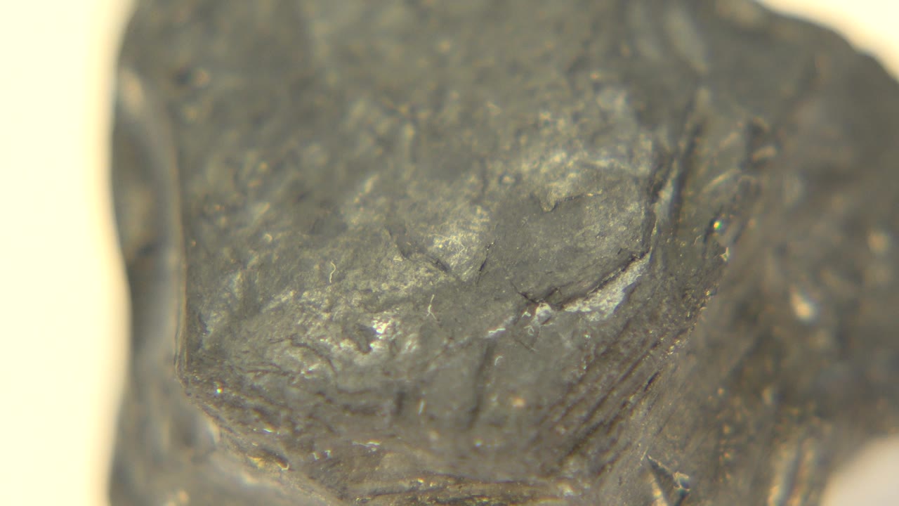 Close macro frame of a dark graphite rock in Westminster, Colorado, with glossy gray facets and chipped surface texture against a bright off-white background, no visible people.