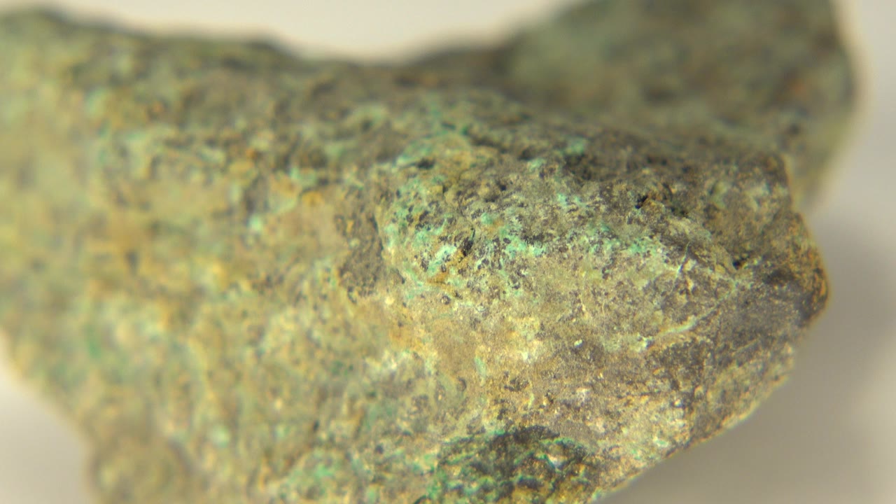 Opening close-up of a rough green, tan, and gray mineral-stained rock in Westminster, Colorado, with a soft pale background and no visible people.