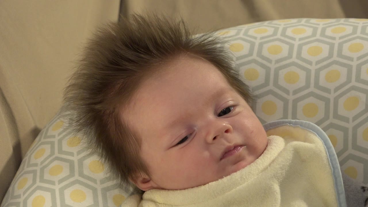 At 0:01, the baby remains swaddled against the patterned pillow, with a sleepy face and eyes slightly more open as he looks near the camera.