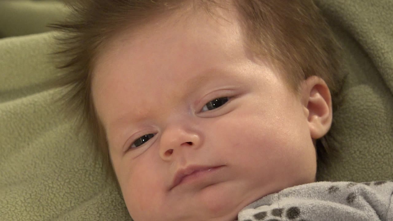 At 0:00, a fair-skinned baby with dark hair lies on a green cushion in Westminster, Colorado, with the face angled slightly left in a tight close-up.