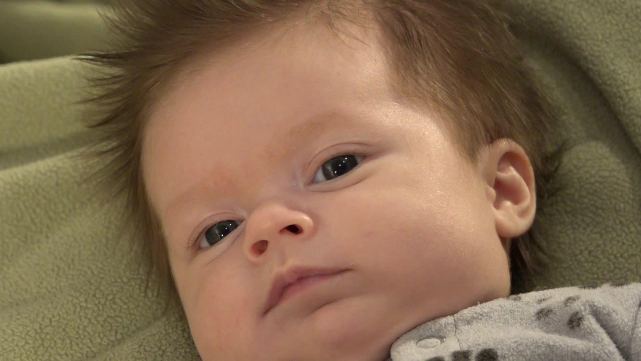 The baby remains close to the lens on green bedding in Westminster, Colorado, with one eye more open and the gray patterned clothing visible at the lower edge.