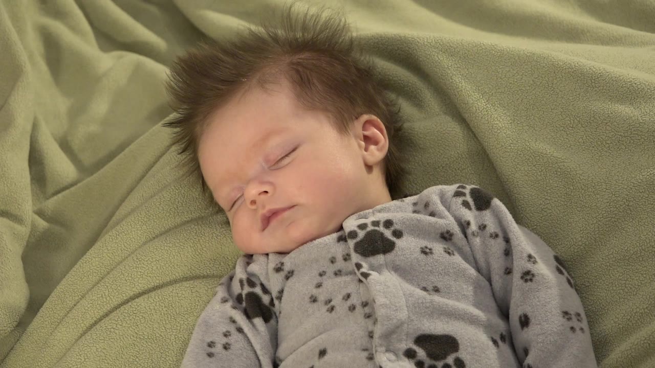 At 0:01, the baby's face remains turned slightly left on the green blanket, with spiky brown hair and the patterned grey pajama collar visible.