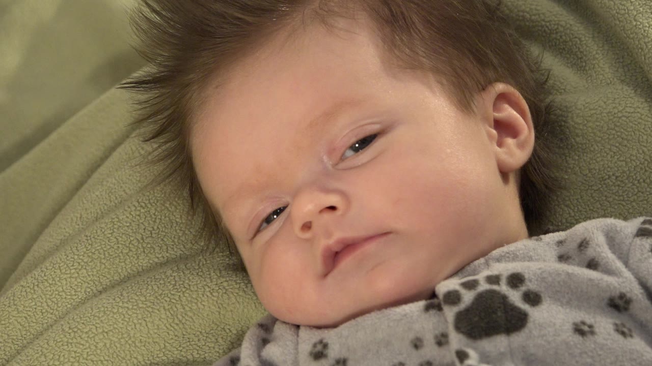At 0:00, a baby boy in Westminster, Colorado lies on a textured green cushion with his eyes partly open and a gray paw-print fleece visible at the lower edge.