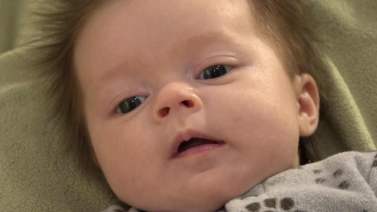 At 0:00, a blond baby with dark eyes lies on green fabric in Westminster, Colorado, looking toward the camera with the mouth slightly open.