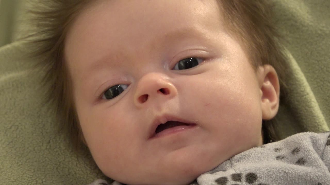 At 0:01, the baby remains close to camera on the green blanket, eyes open and mouth parted as the expression shifts subtly.