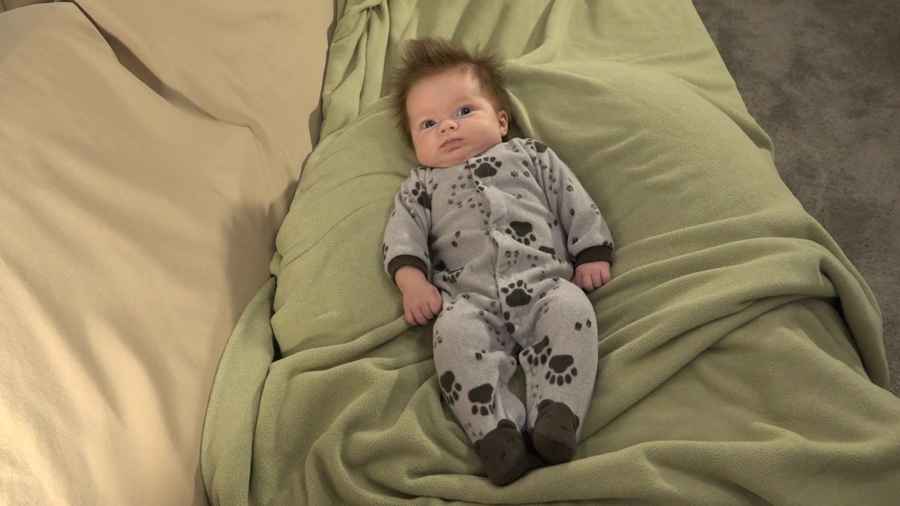 At 0:01, the baby remains centered on the green bedding in Westminster, Colorado, with the cream sheet on the left and the grey floor visible near the top right.