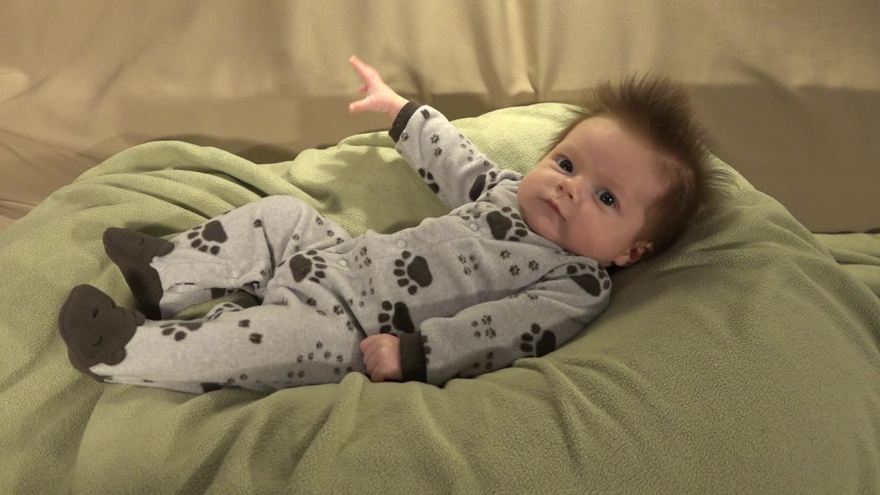 At 0:00, a baby in gray puppy-print pajamas lies on a pale green blanket in Westminster, Colorado, with one arm raised and beige bedding behind.