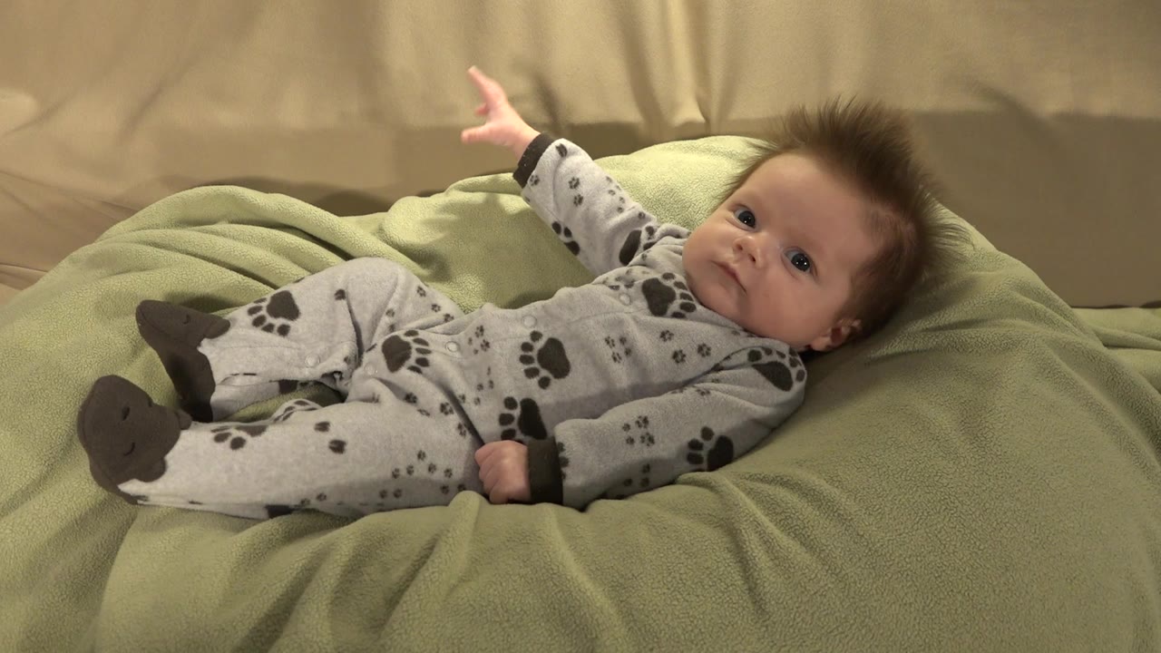 At 0:01, the baby remains reclined on the green blanket in Westminster, Colorado, with an arm stretched above the head and feet angled toward the camera.