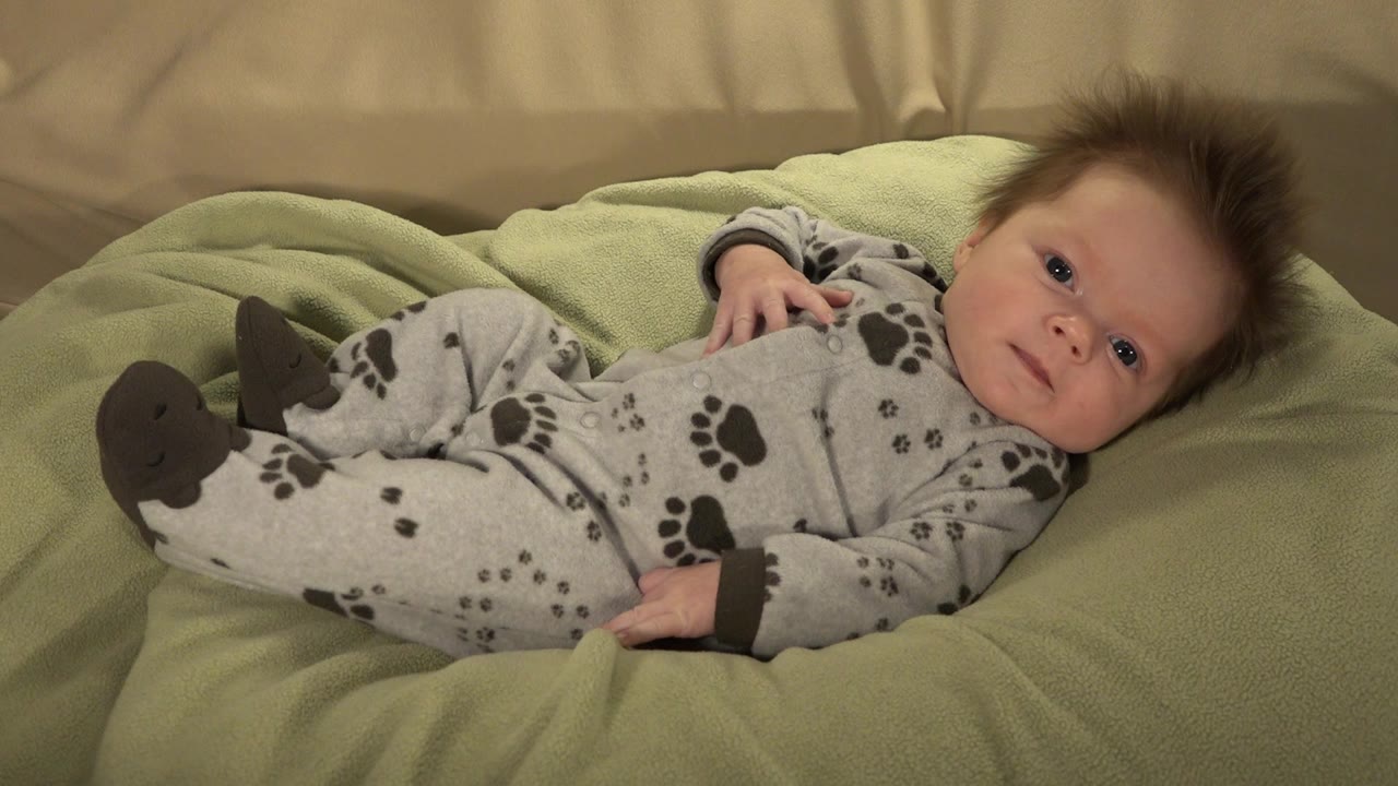 One second in, the baby remains reclined on the green cushion in Westminster, Colorado, with eyes open and one small hand lifted near the shoulder.
