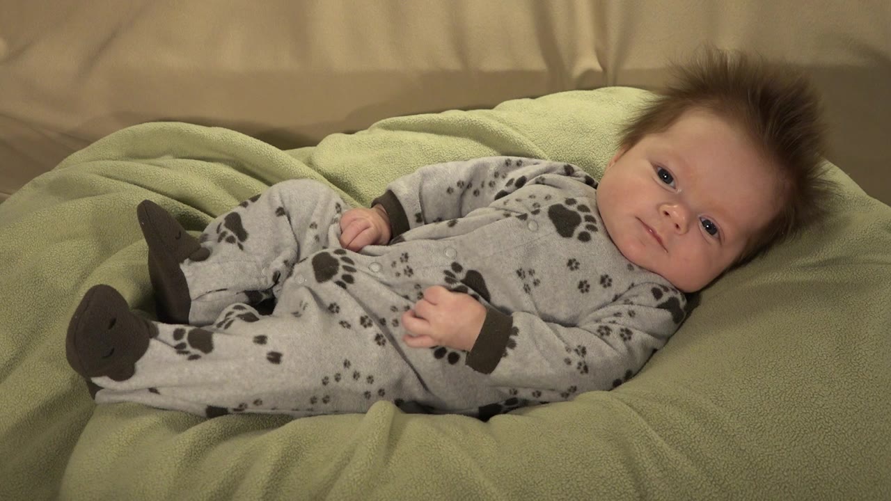 At 0:01 in Westminster, Colorado, the baby looks toward the camera while lying on the green blanket in the gray sleeper.