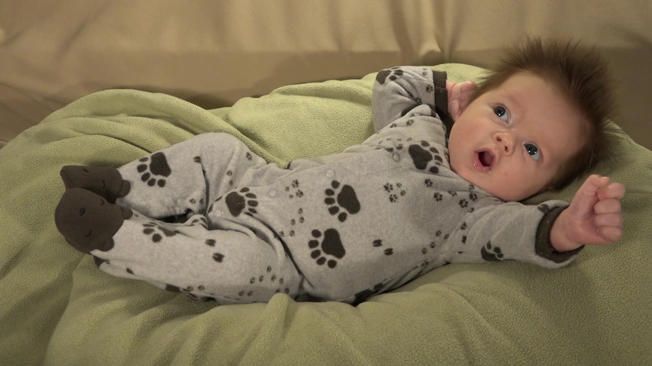 In Westminster, Colorado, the baby remains on the green pillow with eyes turned upward, mouth open, and a small fist lifted near the right side of the frame.