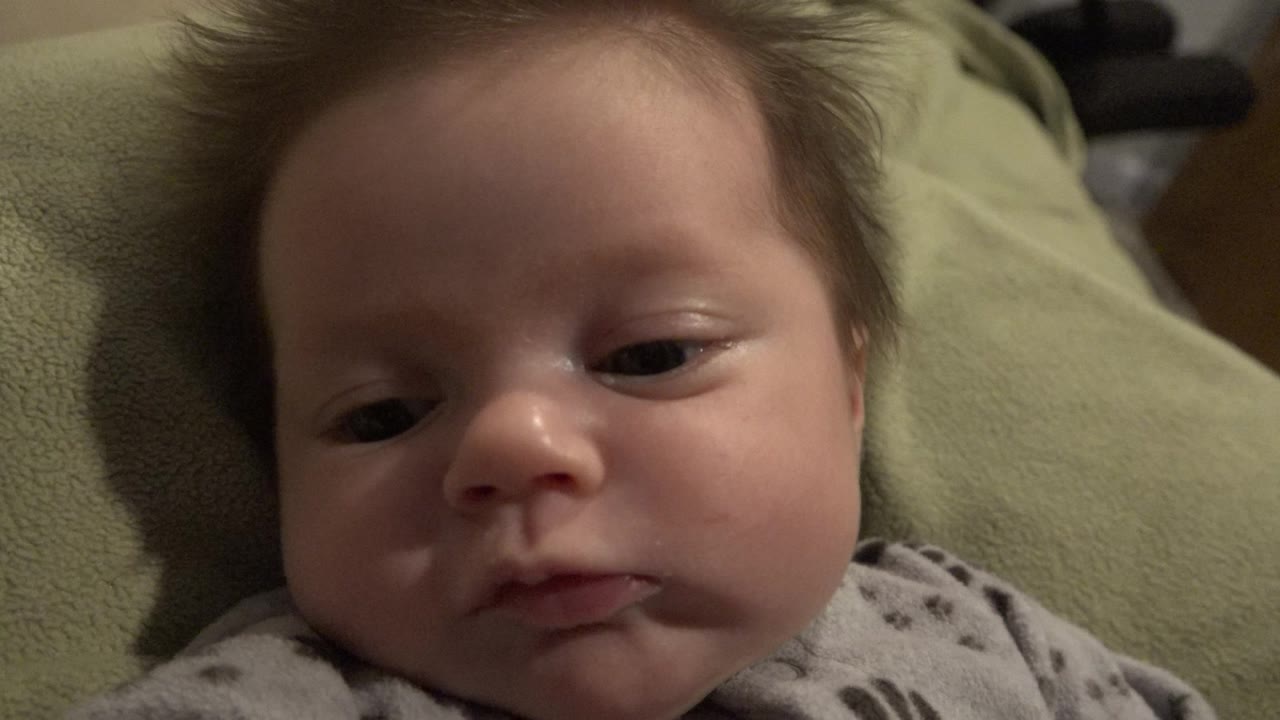 A baby boy in Westminster, Colorado lies close to the camera on a green blanket, his eyes partly open and his gray patterned onesie visible at the bottom of frame.