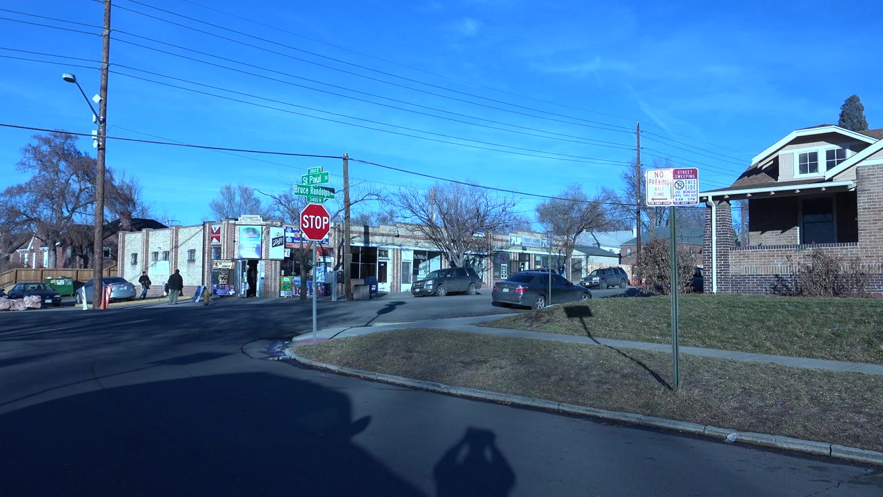 Denver, Colorado street corner at the start of the clip, showing a red stop sign, green street signs, low buildings, parked cars, utility lines, and distant pedestrians.