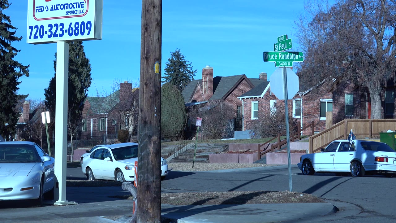 Denver residential street corner with parked cars, brick houses, green street signs, a utility pole, a business sign, and no clearly visible people at the opening moment.
