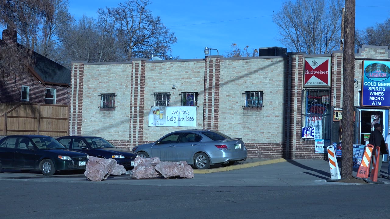 Denver, Colorado storefront row seen across a paved street, with parked sedans, large rocks along the curb, bare trees, branded signs, and no visible people in this opening frame.