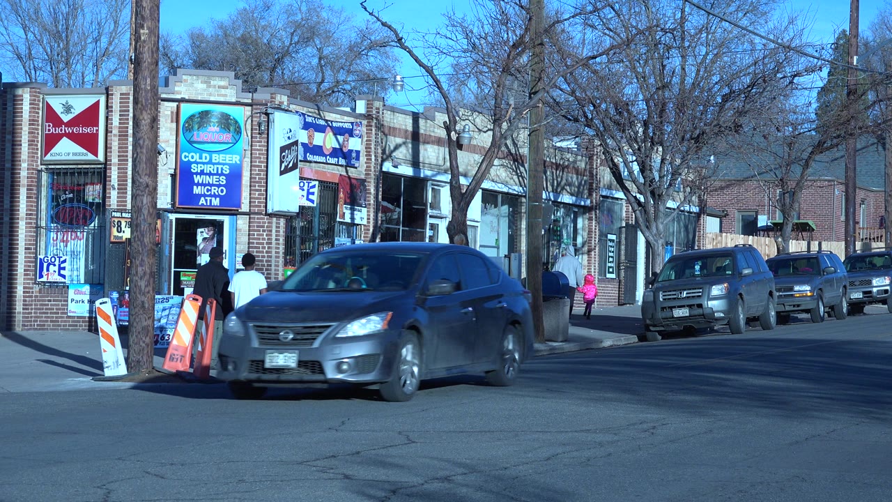 Denver street corner in 2015 with storefront advertising, parked vehicles, leafless trees, and a dark sedan crossing the foreground as people stand near the shop entrance.