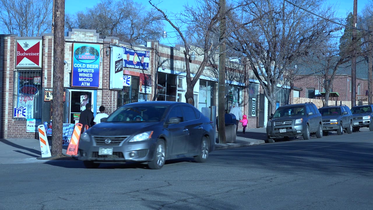 Denver storefront block with a dark sedan in the foreground, a person in a white shirt near the entrance, and parked vehicles lining the curb under bare trees.