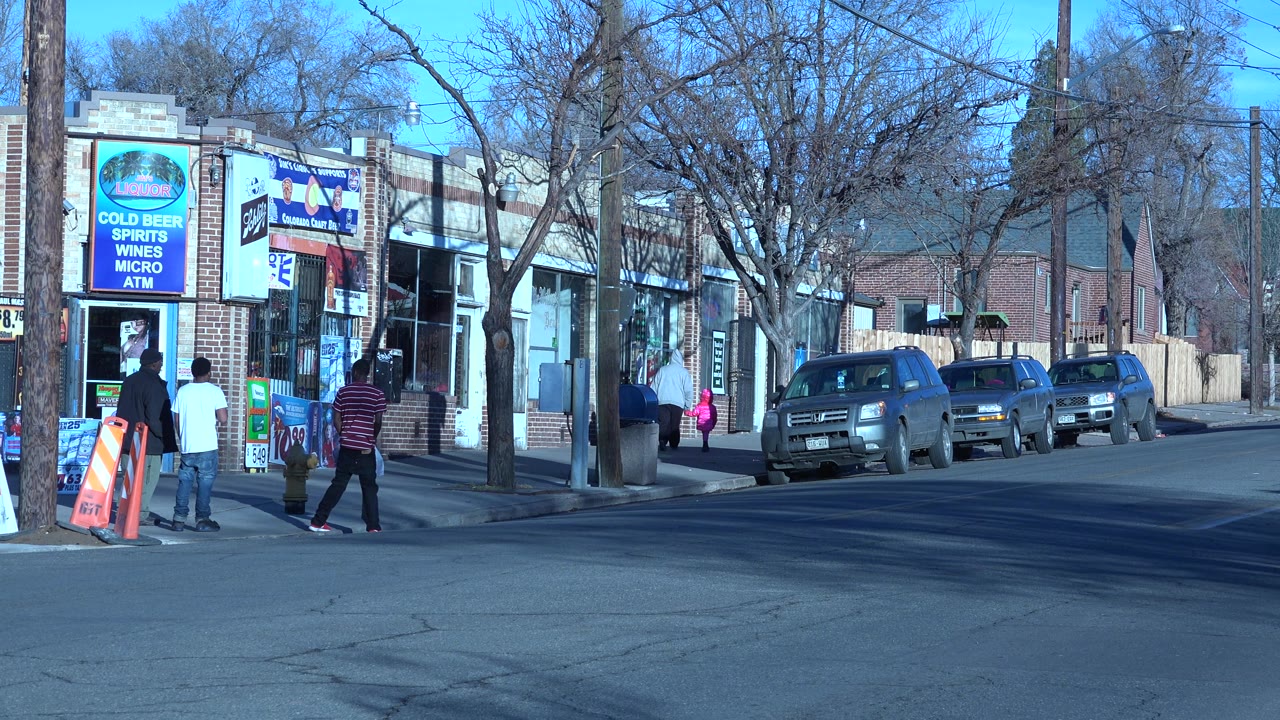 Pedestrians walk past a corner store with parked vehicles along the street.