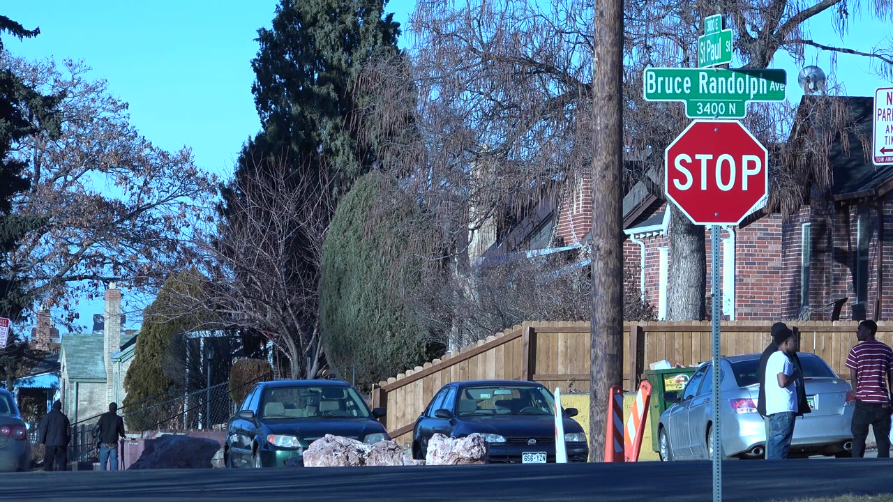 The Denver intersection remains framed around the stop sign, with two people beside a silver car and brick homes or storefronts behind them in bright sun.