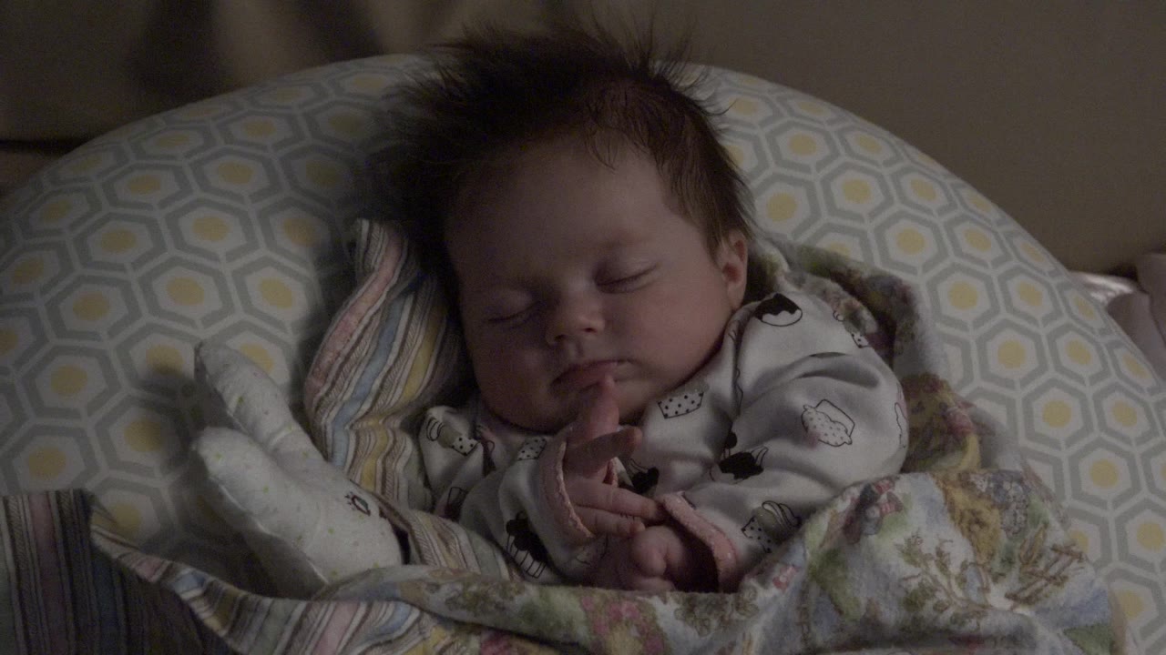 A sleeping baby lies on layered patterned pillows and blankets in Westminster, Colorado, with the face and hands visible in dim indoor light.