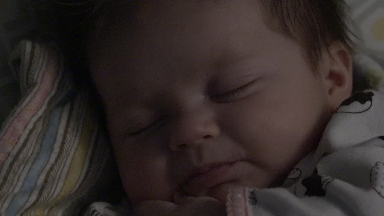 The baby remains asleep in a close Westminster, Colorado home-movie frame, with the face filling most of the image and the striped pillow visible along one side.