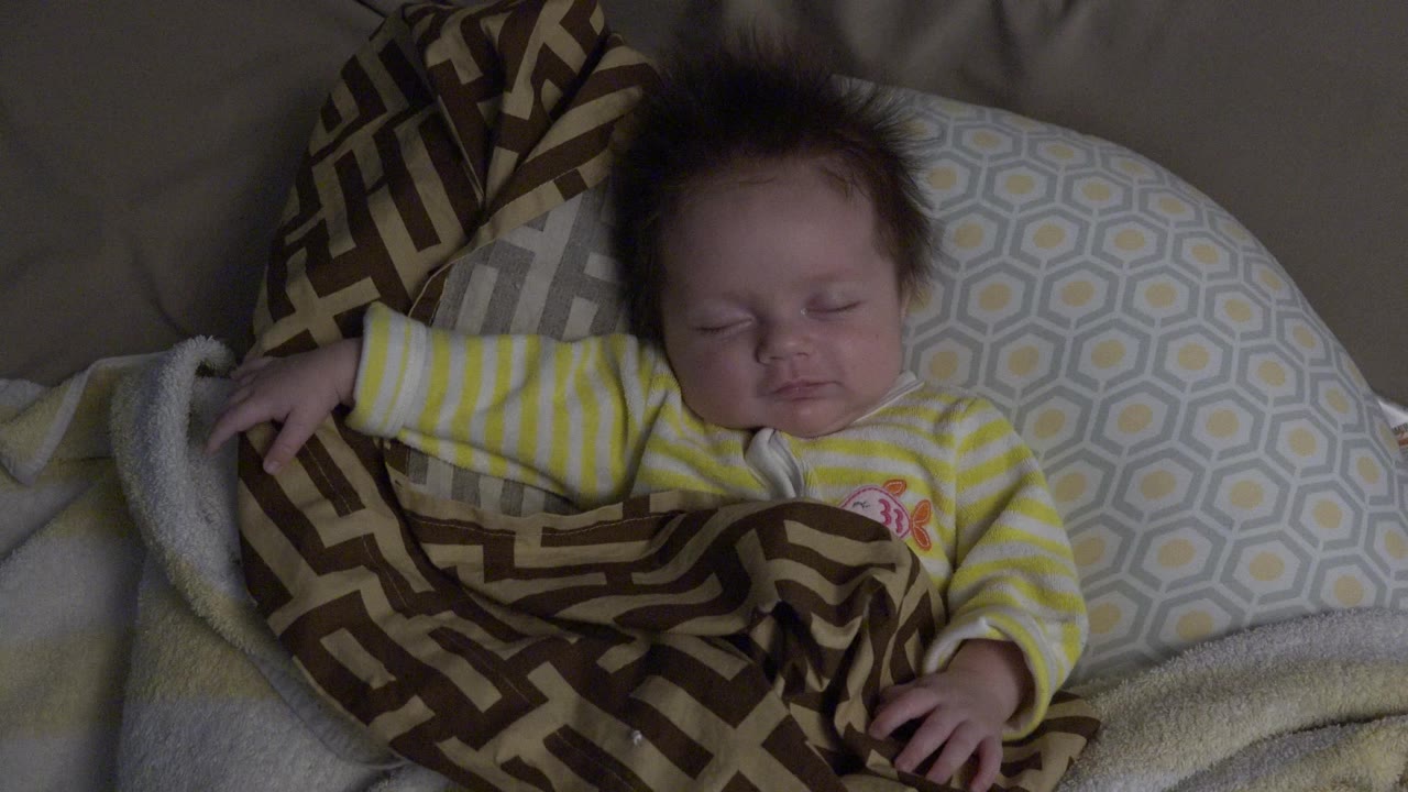At 0:01, the baby remains asleep on the pillow arrangement, with the brown patterned blanket covering the lower body and the yellow sleeve stretched outward.
