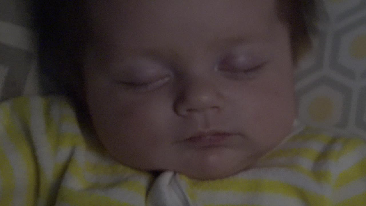 At 0:01, the sleeping infant remains tightly framed, with the striped sleeve and gray patterned pillow more visible beside the face in Westminster, Colorado.