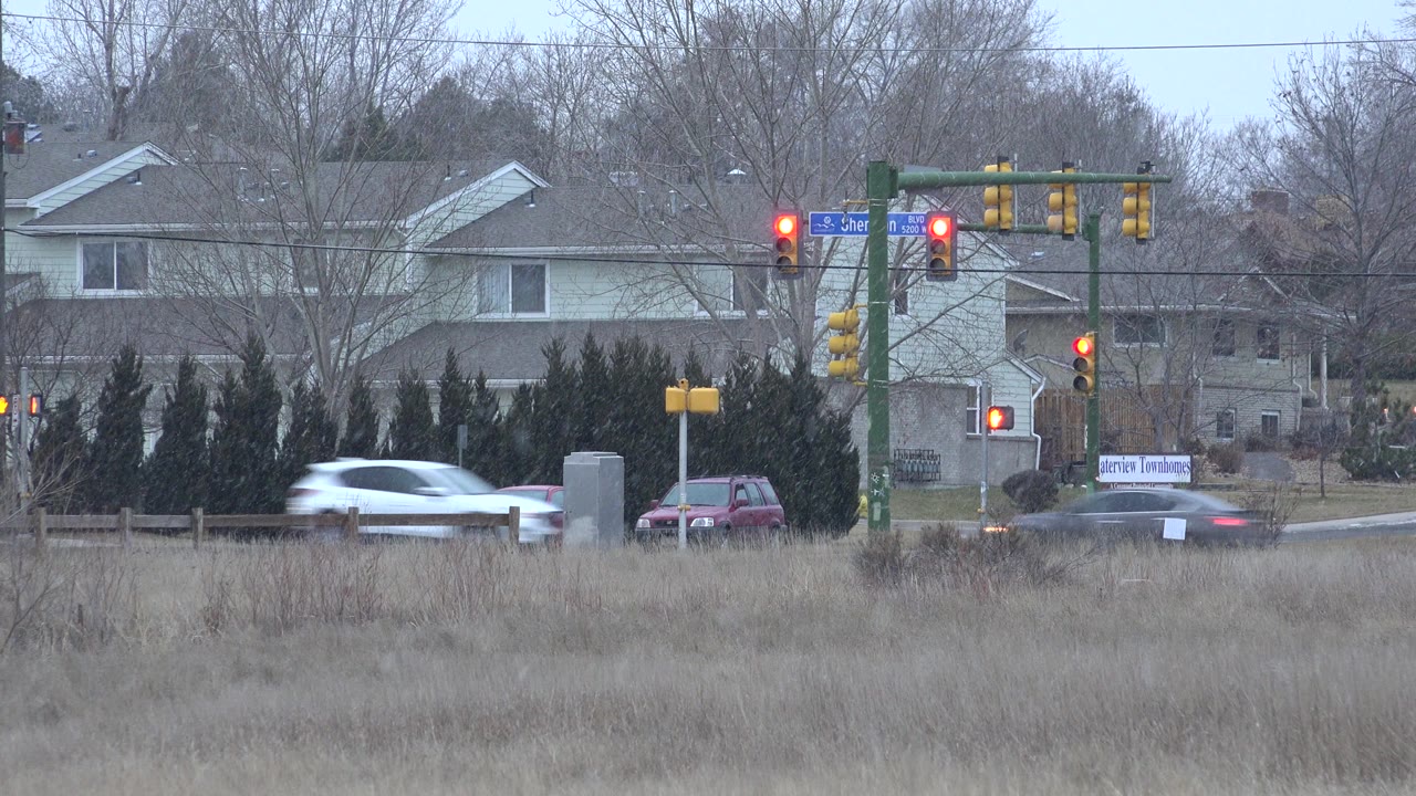A white car crosses the Westminster, Colorado road scene while red lights hold at the intersection, with bare trees, shrubs, private homes, and no visible people.
