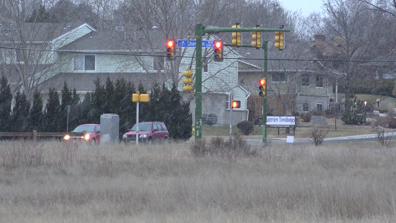 Westminster, Colorado junction view with red signal lights, a red car and other vehicles near the road, pale houses in the background, dry grass below, and no visible people.