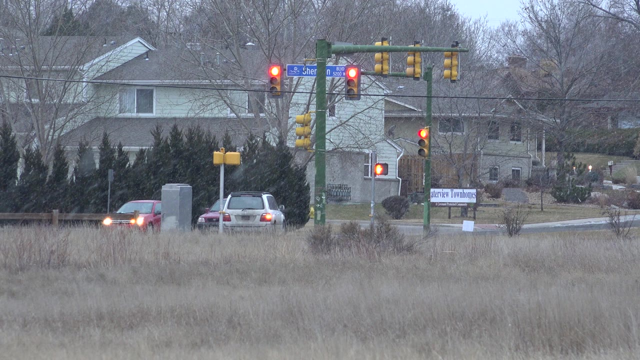 A red car and a light-colored car sit near the Westminster, Colorado intersection as multiple red signals glow above the road, with residential buildings and leafless trees in the background.
