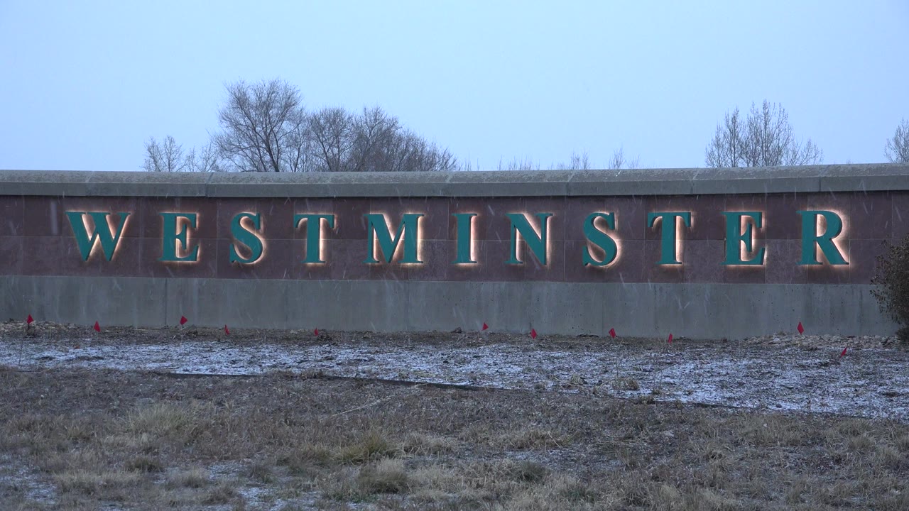 Wide view of the WESTMINSTER sign in Westminster, Colorado, with patchy snow on grass below, bare trees above the wall, and no visible people.