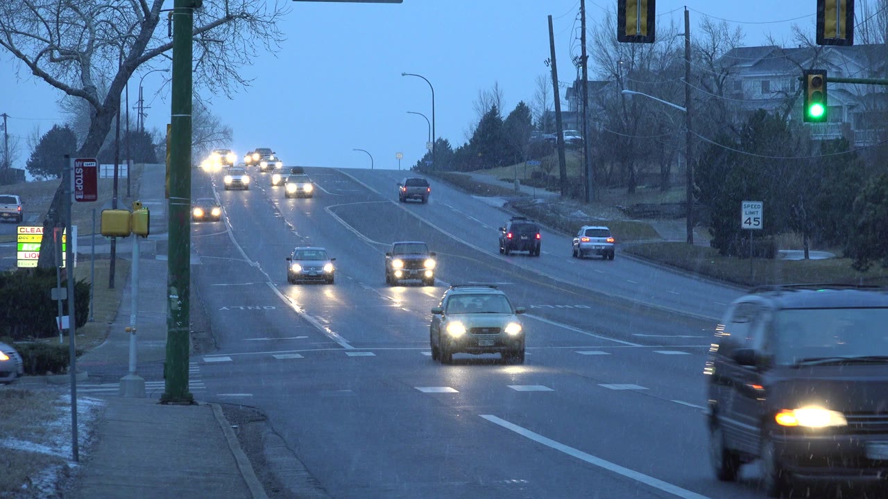 Cars with headlights approach a snowy suburban intersection under a green traffic light.