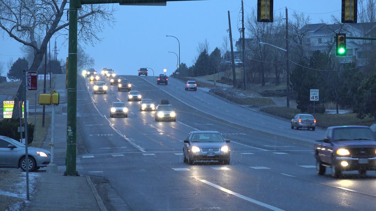 At 0:00 in Westminster, Colorado, cars with headlights approach a green traffic light on a wet multi-lane avenue, with bare trees, roadside signs, and distant homes in view and no visible people.