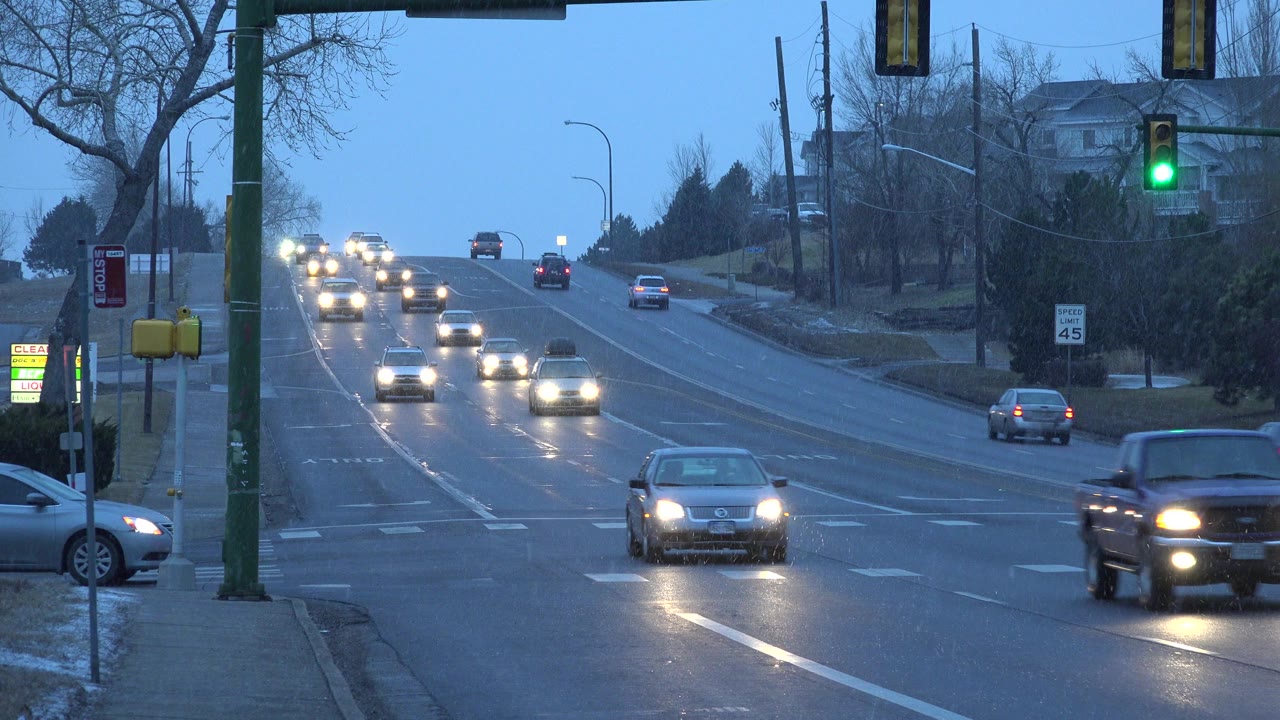 At 0:01 in Westminster, Colorado, a dark pickup passes across the lower right foreground as headlight traffic approaches on the wet avenue under the green signal, with no visible people.