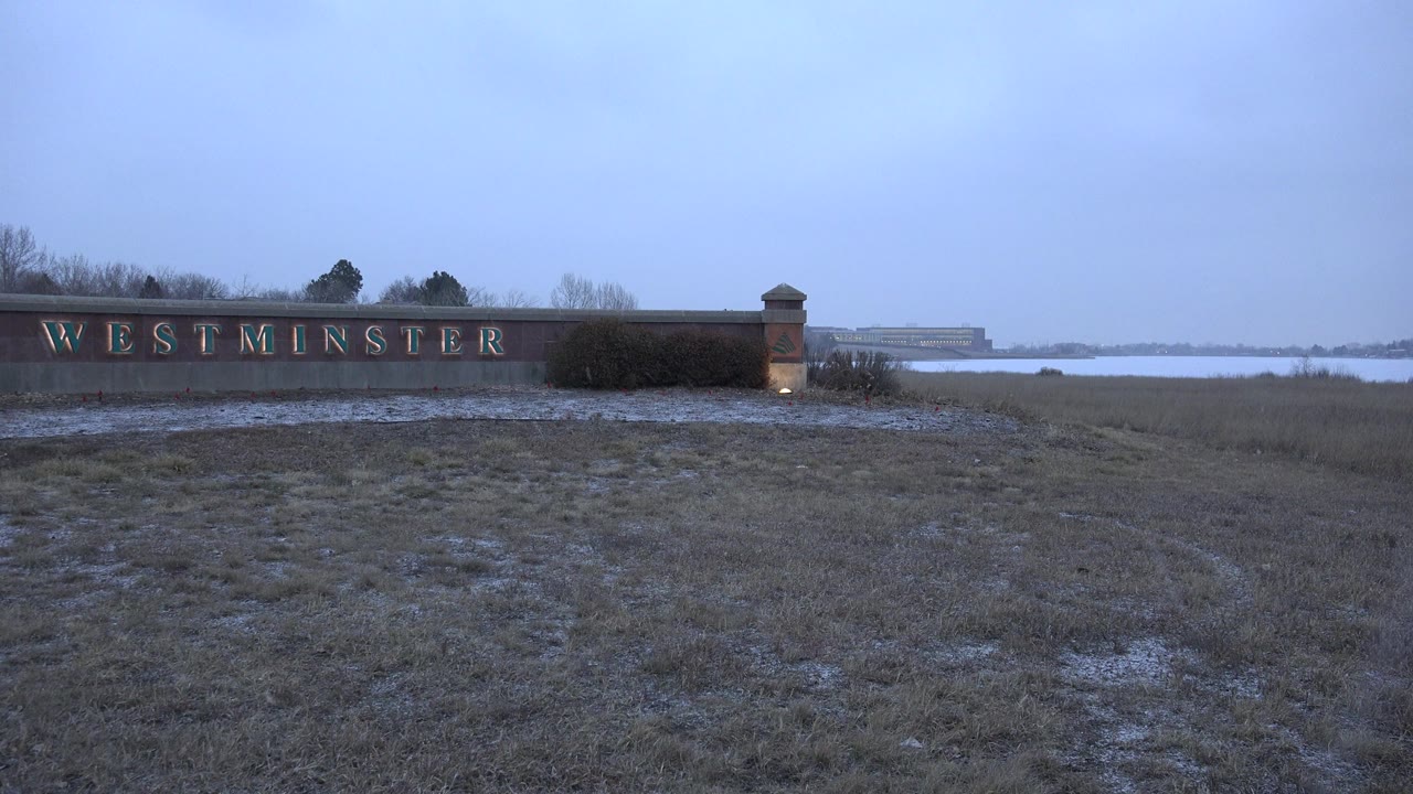 The Westminster sign remains left of center with frosted grass in the foreground and a broad water surface behind it in Westminster, Colorado, with no visible people.