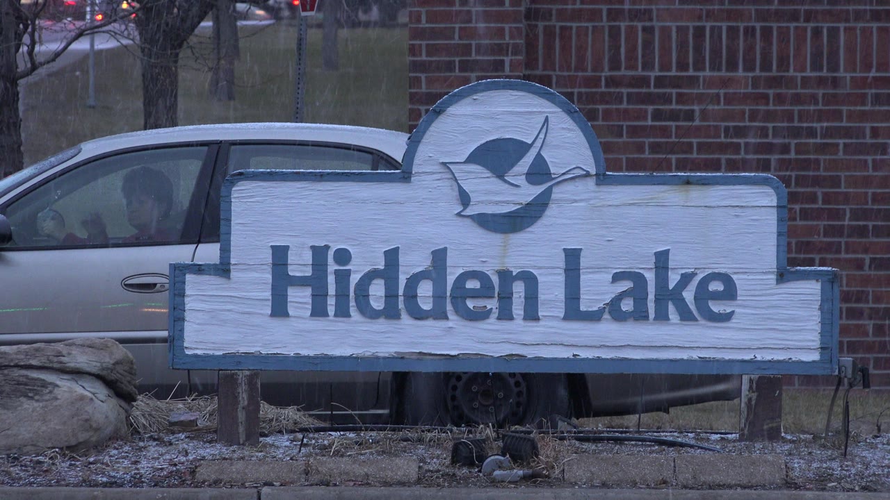 At 0:01, the Hidden Lake sign remains centered, with the silver car more visible behind the left side and the brick wall filling the background in Westminster, Colorado.