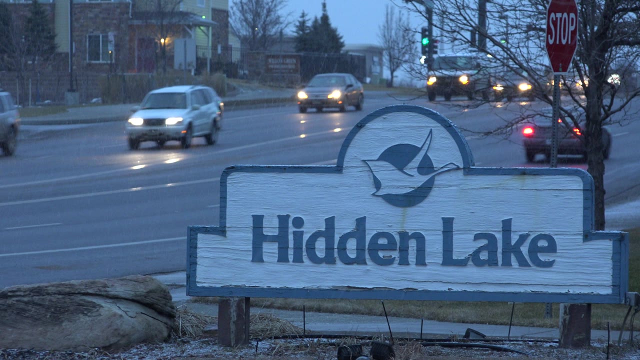 The Hidden Lake sign remains centered in Westminster, Colorado, as headlights and passing cars move through the wet intersection area behind it, with no visible people.