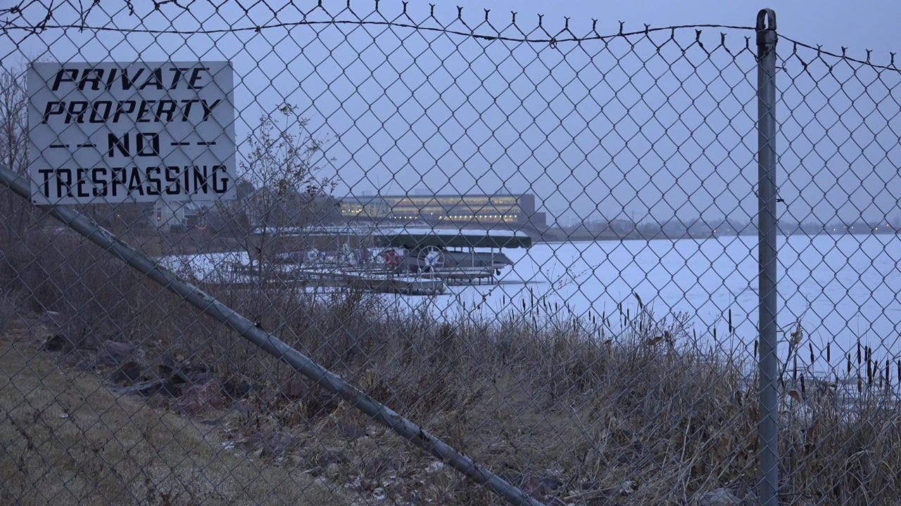 In Westminster, Colorado, a chain-link fence and PRIVATE PROPERTY NO TRESPASSING sign frame a lake edge with a metal grill or picnic fixture beyond it and no visible people.