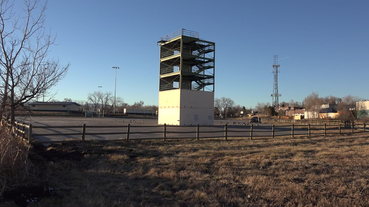 A Westminster, Colorado training tower stands beside an open paved area with a wooden fence, dry grass, bare trees, and no visible people at the start of the clip.
