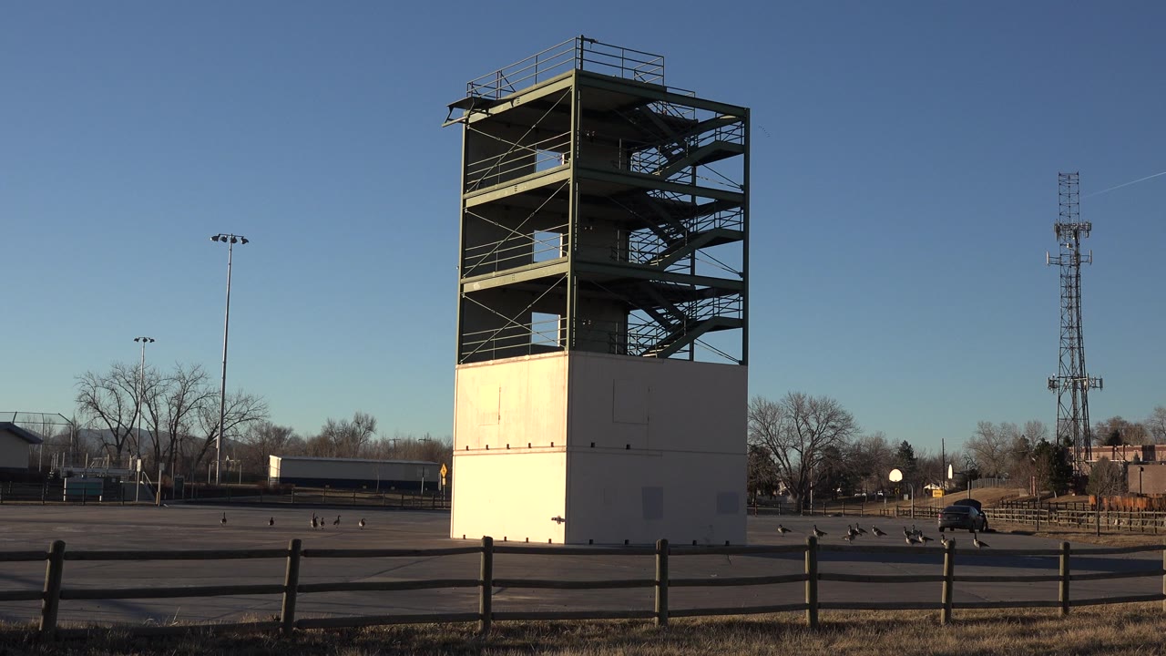 Wide view of an unfinished stair tower in Westminster, Colorado, with a low fence in the foreground, distant trees and utility structures behind it, and no visible people.